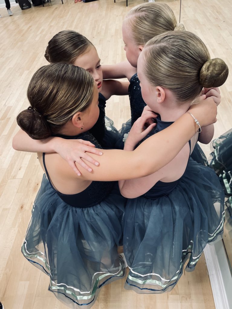 Four young girls dressed in blue and black dance costumes huddle together in a circle at a dance studio
