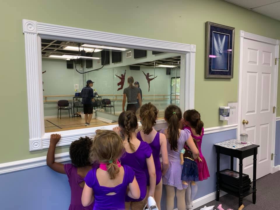 A group of 6 children are seen from behind gathered together and looking through a viewing window to a dance class in progress
